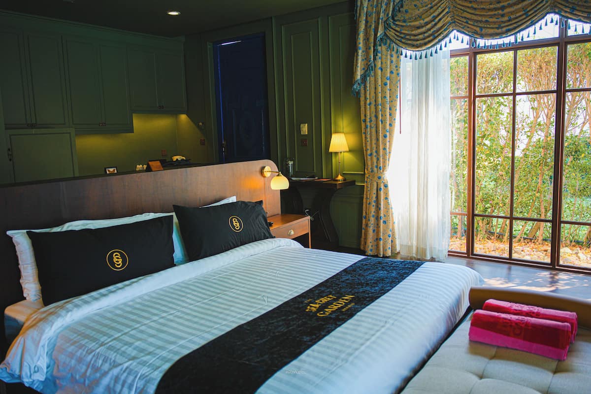 A hotel room with a large bed, a desk with a lamp, a window with curtains, and a view of trees.