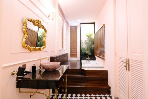 Un pasillo estrecho con suelo a cuadros blanco y negro, un tocador con lavamanos rosa y detalles dorados, y una escalera que conduce a un cuarto con bañera y un ventanal grande.