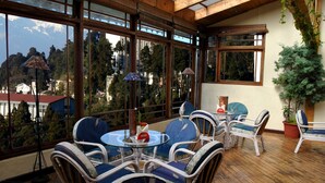 A sunlit patio with wicker furniture and a glass table, overlooking a mountainous landscape. - Central Heritage Resort & Spa, Darjeeling (Darjeeling)