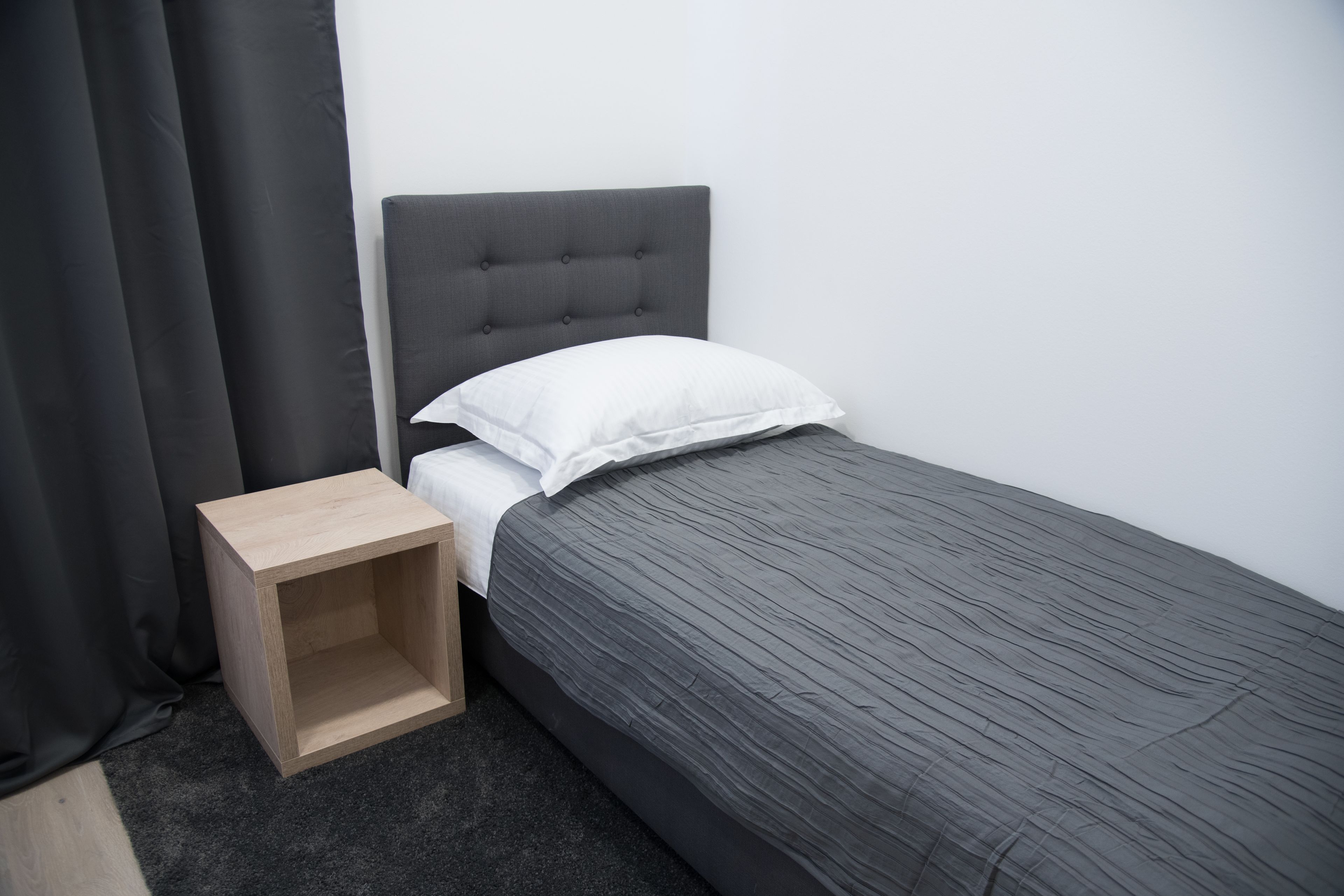 A single bed with a grey headboard and white pillow, a wooden nightstand, and a dark grey carpet.