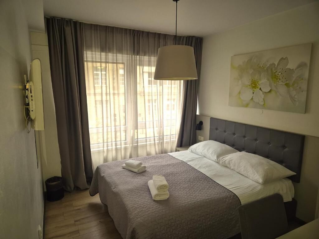 Habitación de hotel con cama, un ventanal con cortinas, un cuadro en la pared y una lámpara en el techo.