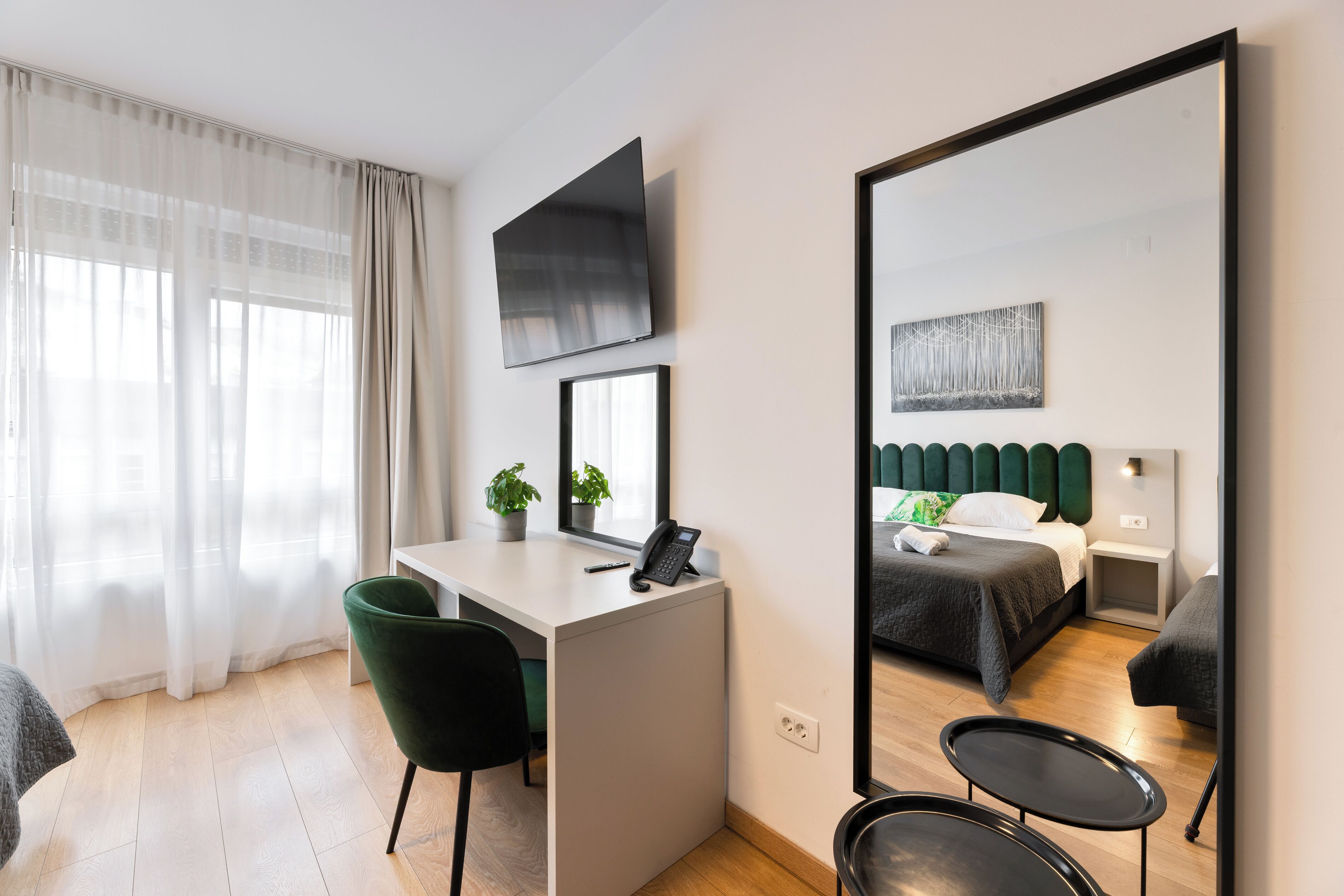 Een moderne hotelkamer met een bureau, stoel, bed, nachtkastje en een grote spiegel.