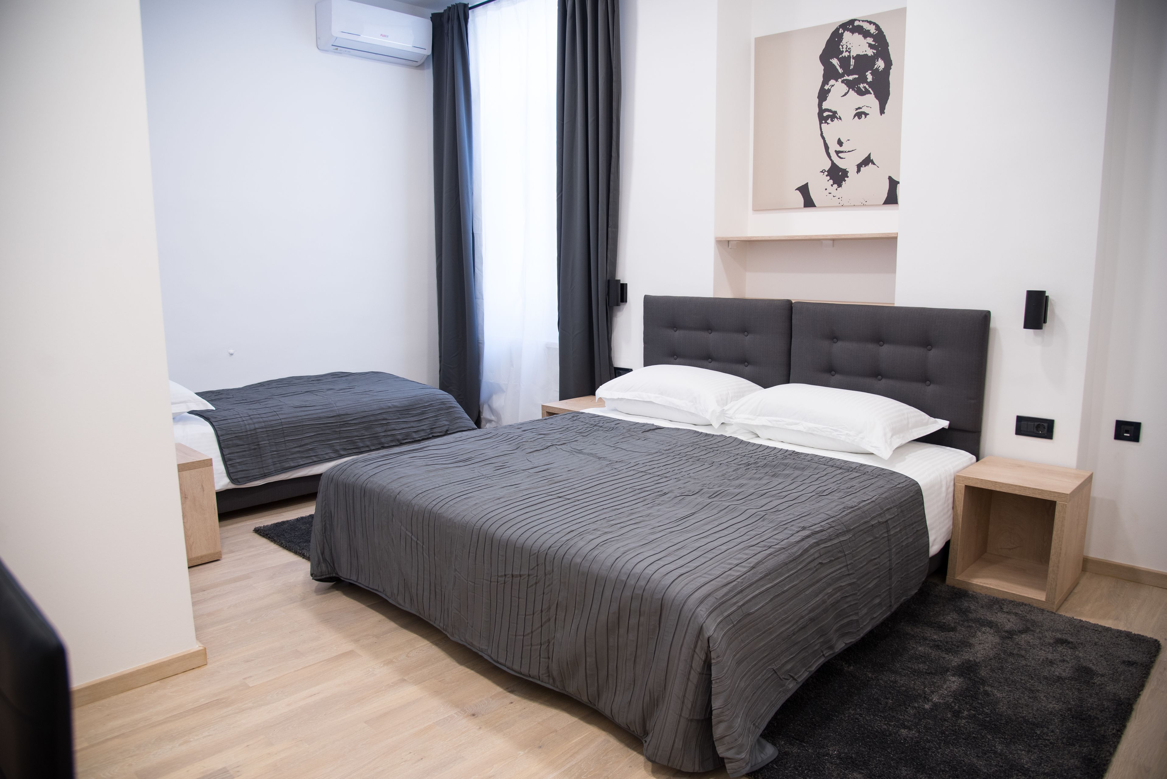 A modern hotel room with a large bed, a smaller bed, a bedside table, a nightstand, a black rug, and a wall-mounted air conditioning unit.