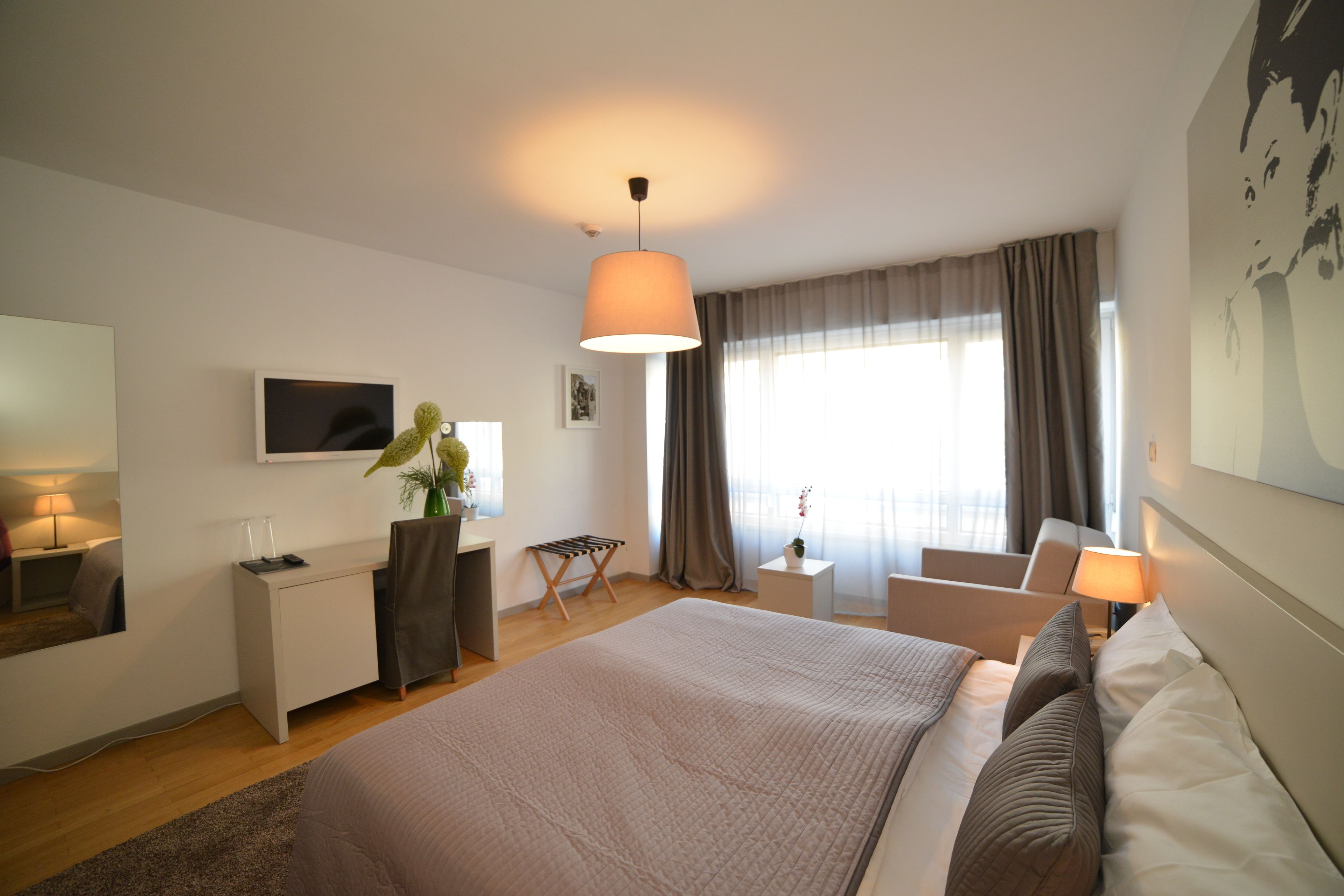 Una habitación de hotel moderna con una cama grande, un escritorio, una silla, un televisor y un ventanal con cortinas.