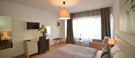 Una habitación de hotel moderna con una cama grande, un escritorio, una silla, un televisor y un ventanal con cortinas.
