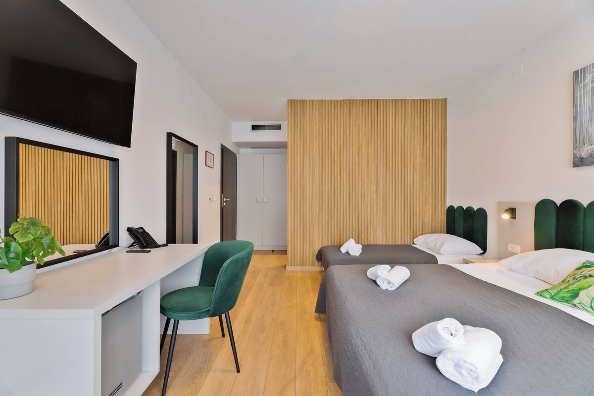 Ein Hotelzimmer mit Schreibtisch, zwei Betten, einem grünen Sessel und einer Topfpflanze.
