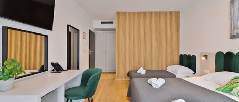 A hotel room with a desk, two beds, a green chair, and a potted plant.