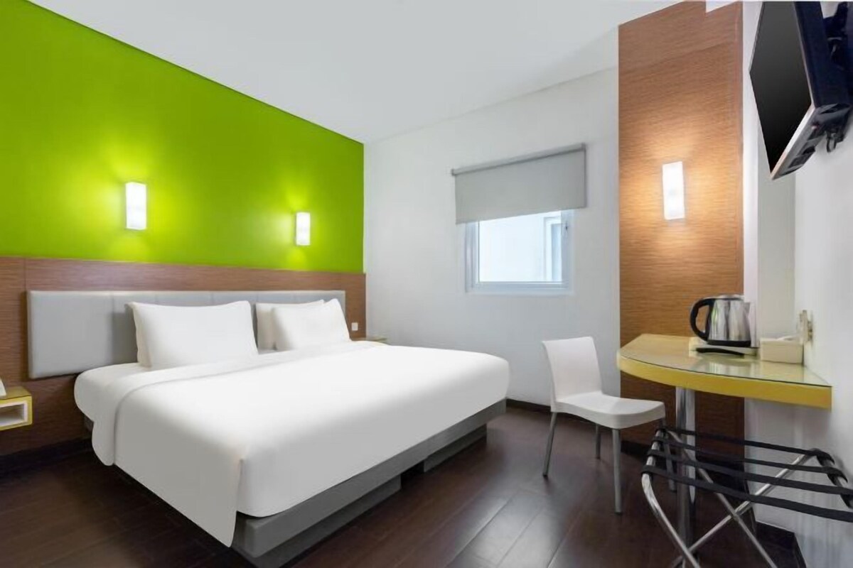 Quarto de hotel com cama, escrivaninha com cadeira, televisão na parede e janela com vista para o exterior.