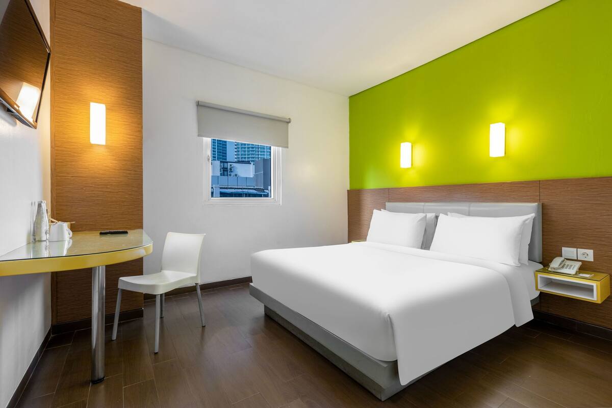 A hotel room with a bed, a small table with a chair, a window, and a wall-mounted phone.