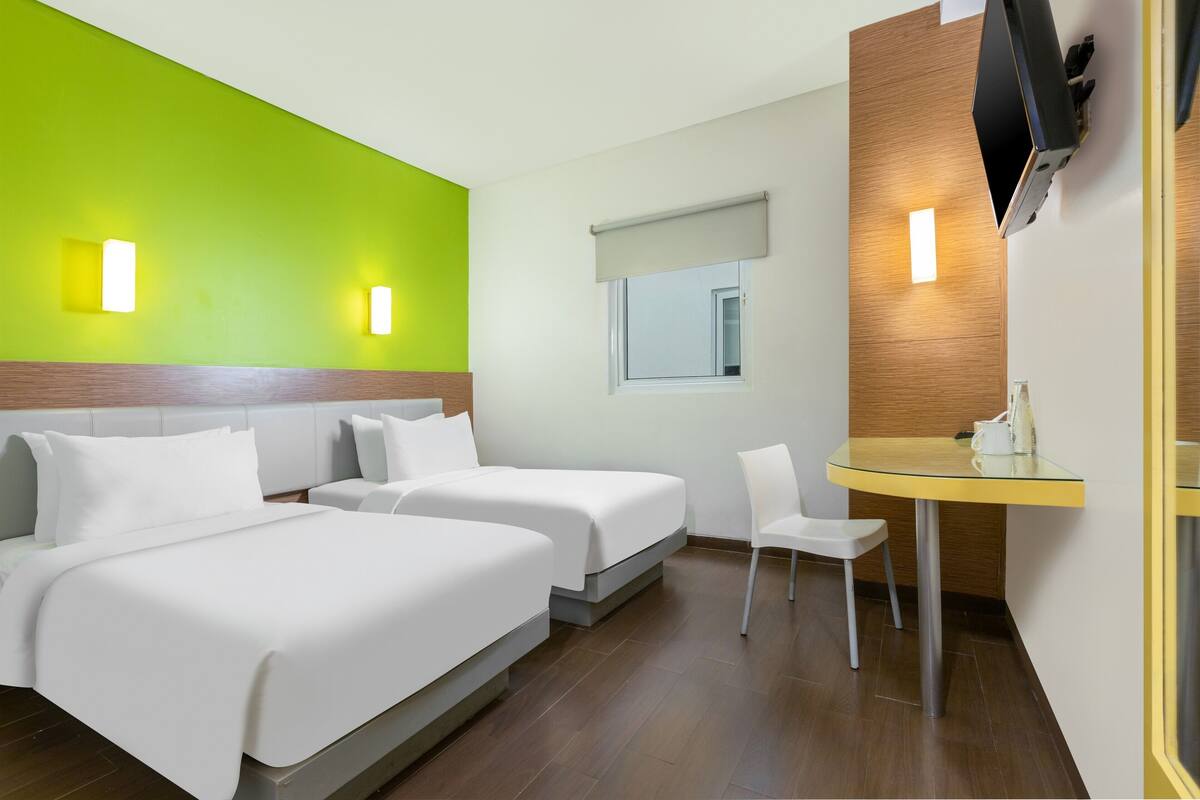 A hotel room with two beds, a small table with a chair, a TV mounted on the wall, and a window with a blind.