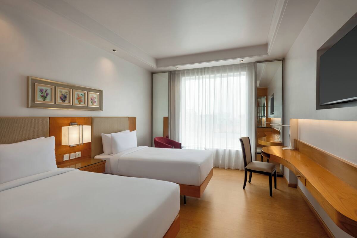 A hotel room with two beds, a desk, a chair, a TV, and a large window with curtains.