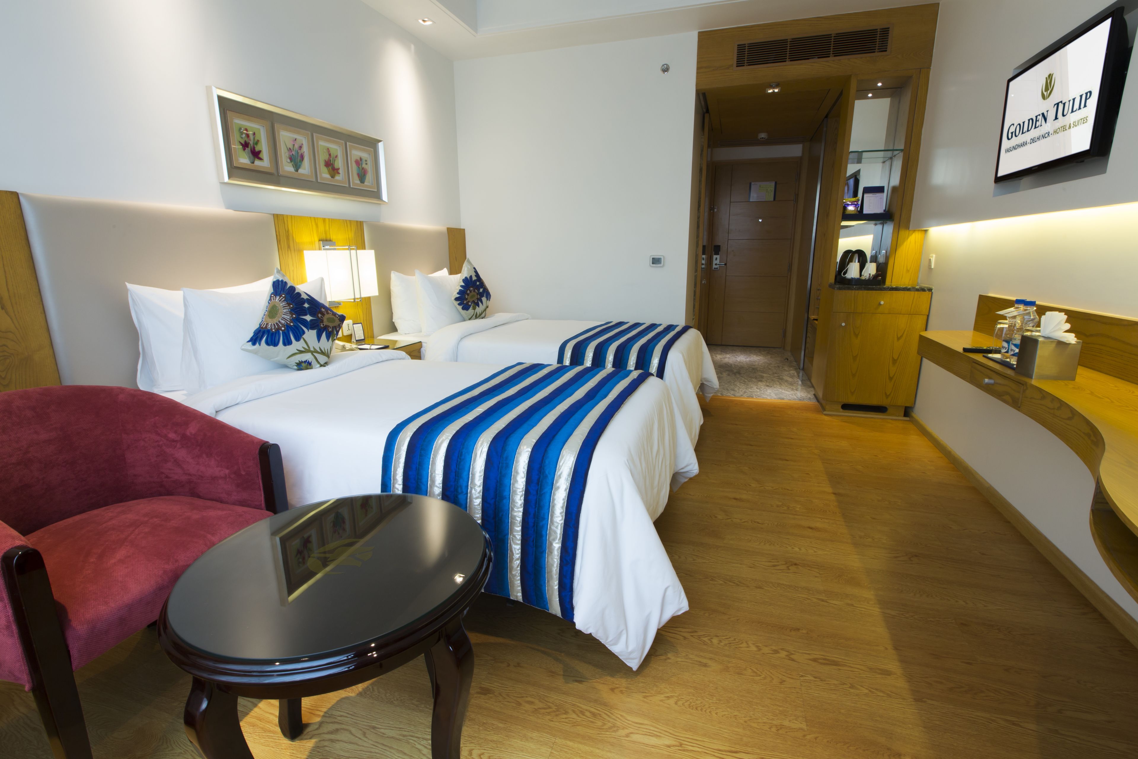 A hotel room with two beds, a red chair, a small table, and a kitchenette.