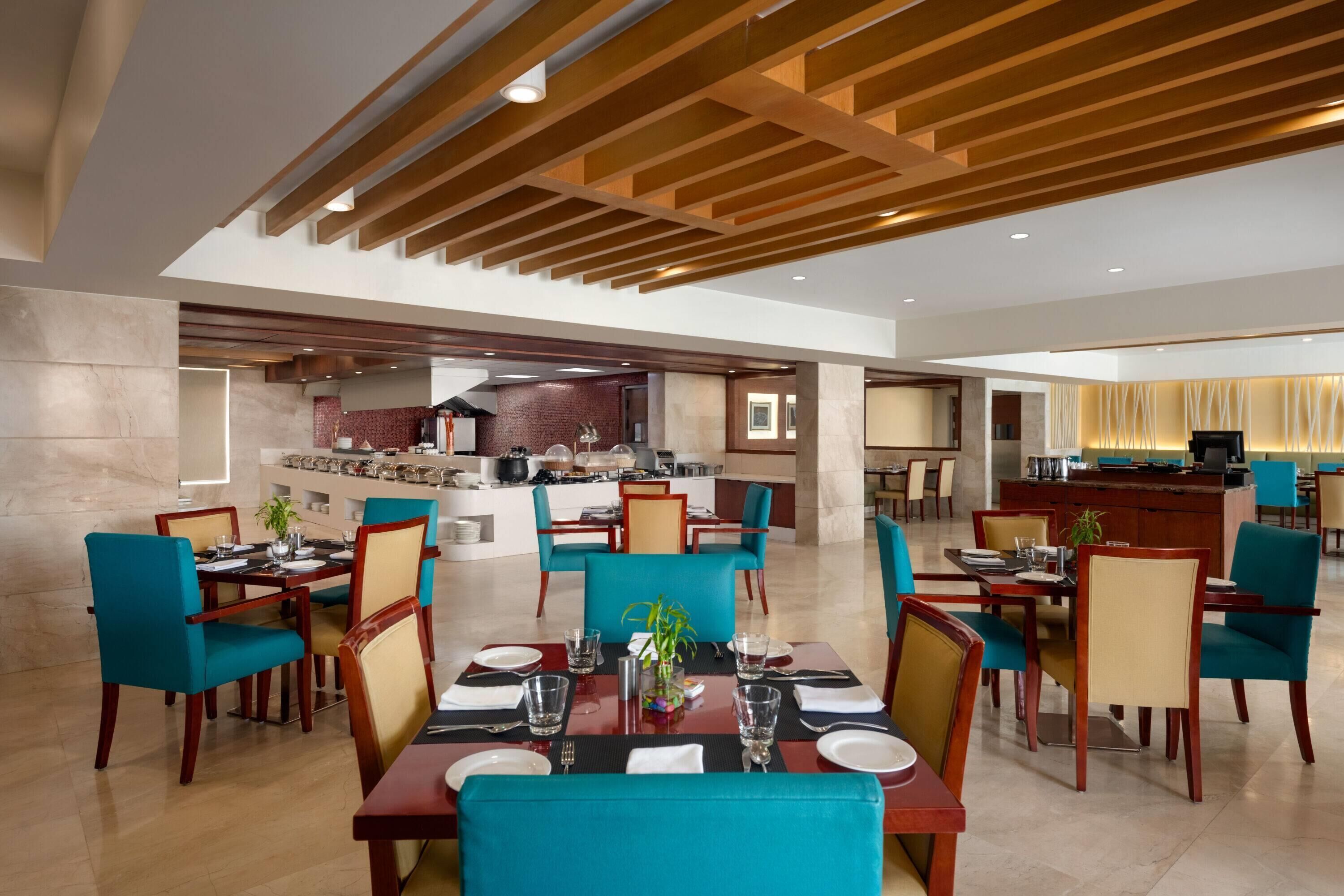 A spacious dining area with multiple tables and chairs, a kitchen in the background, and a modern ceiling design.