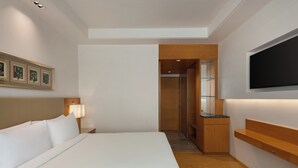 Quarto de hotel com uma cama grande, uma televisão fixada na parede, uma mesa de cabeceira de madeira com abajur e um banco de madeira.