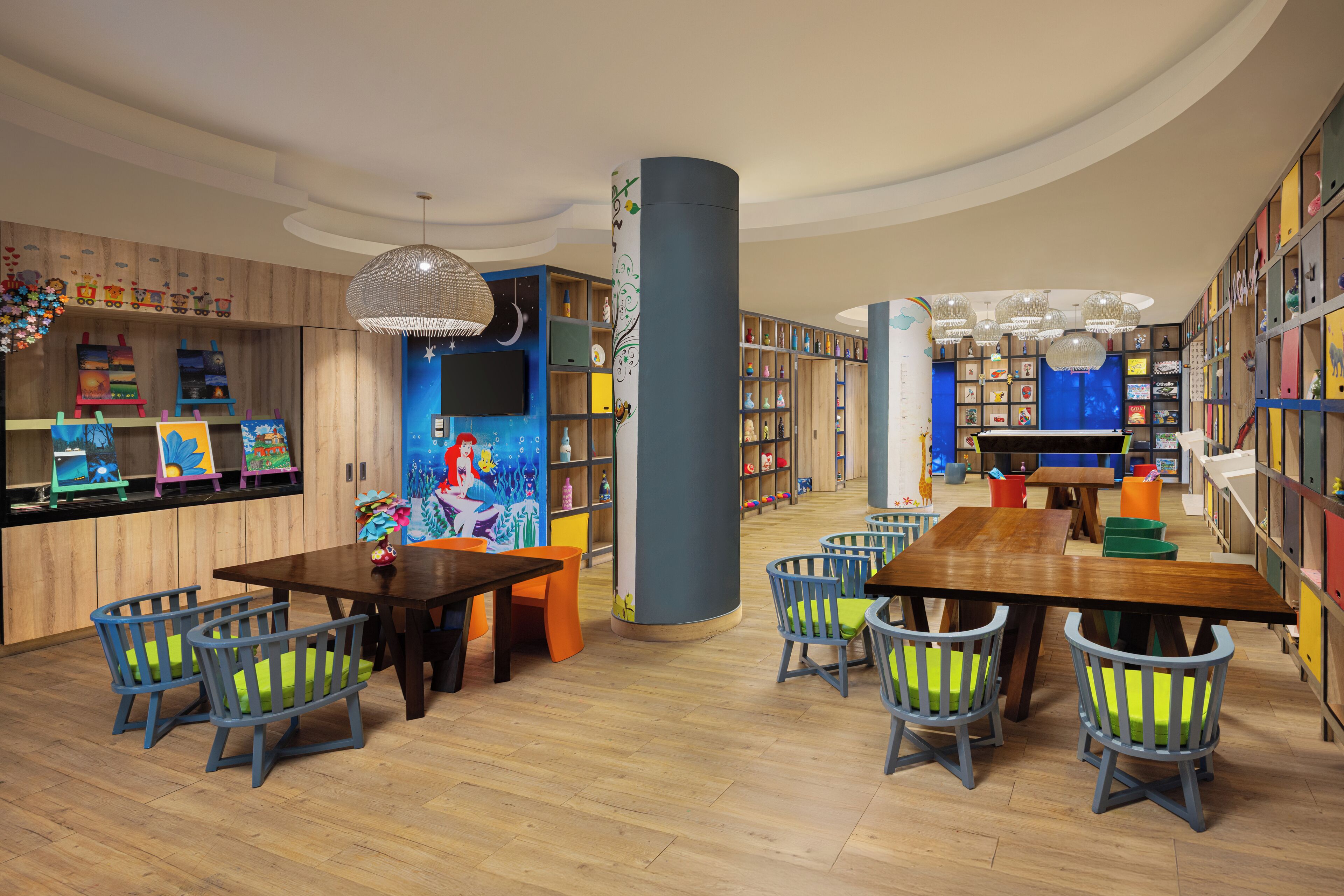 A children's library with wooden floors, a central table, and colorful chairs.