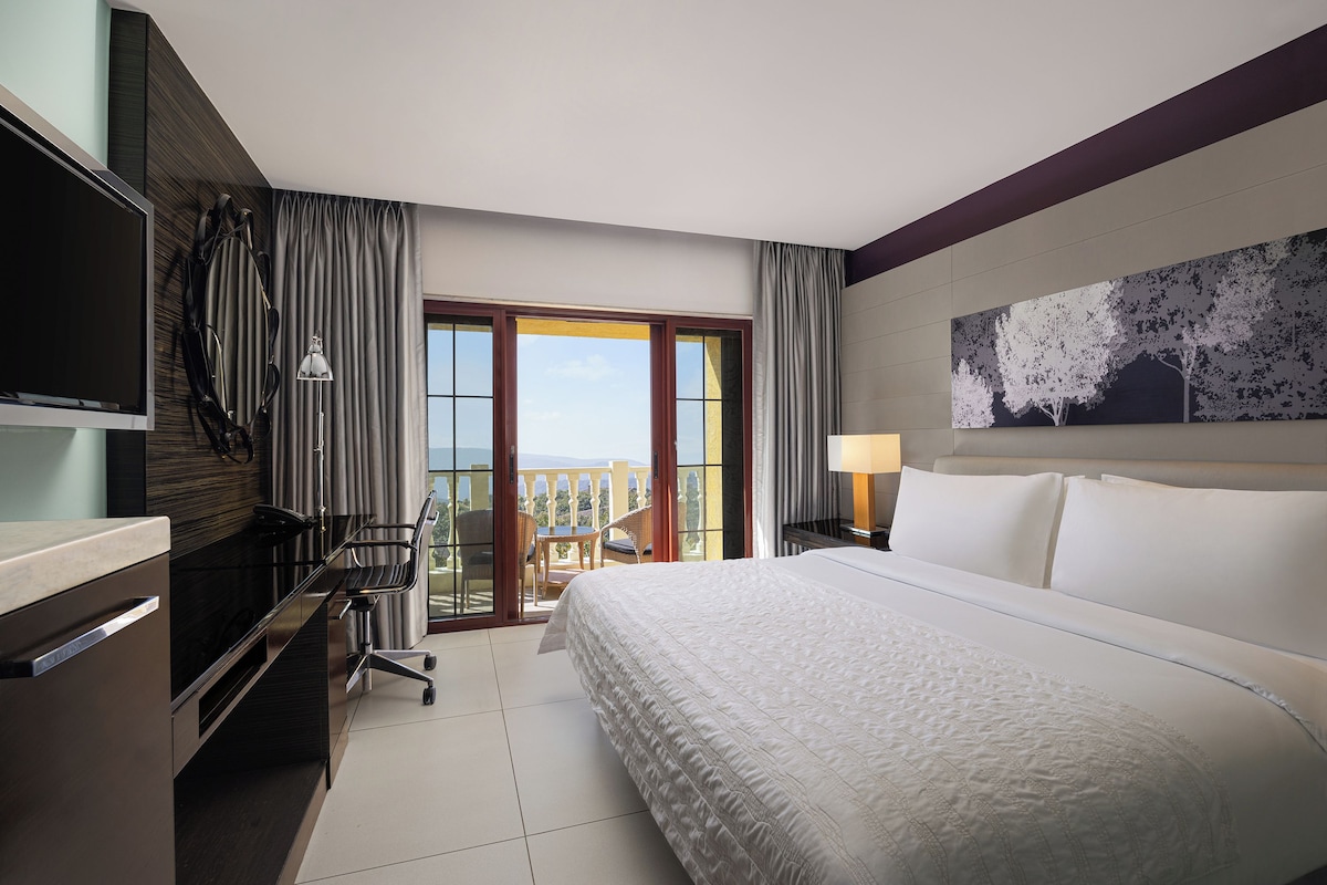Een hotelkamer met een bed, bureau, stoel, televisie, balkon met uitzicht en een schilderij aan de muur.
