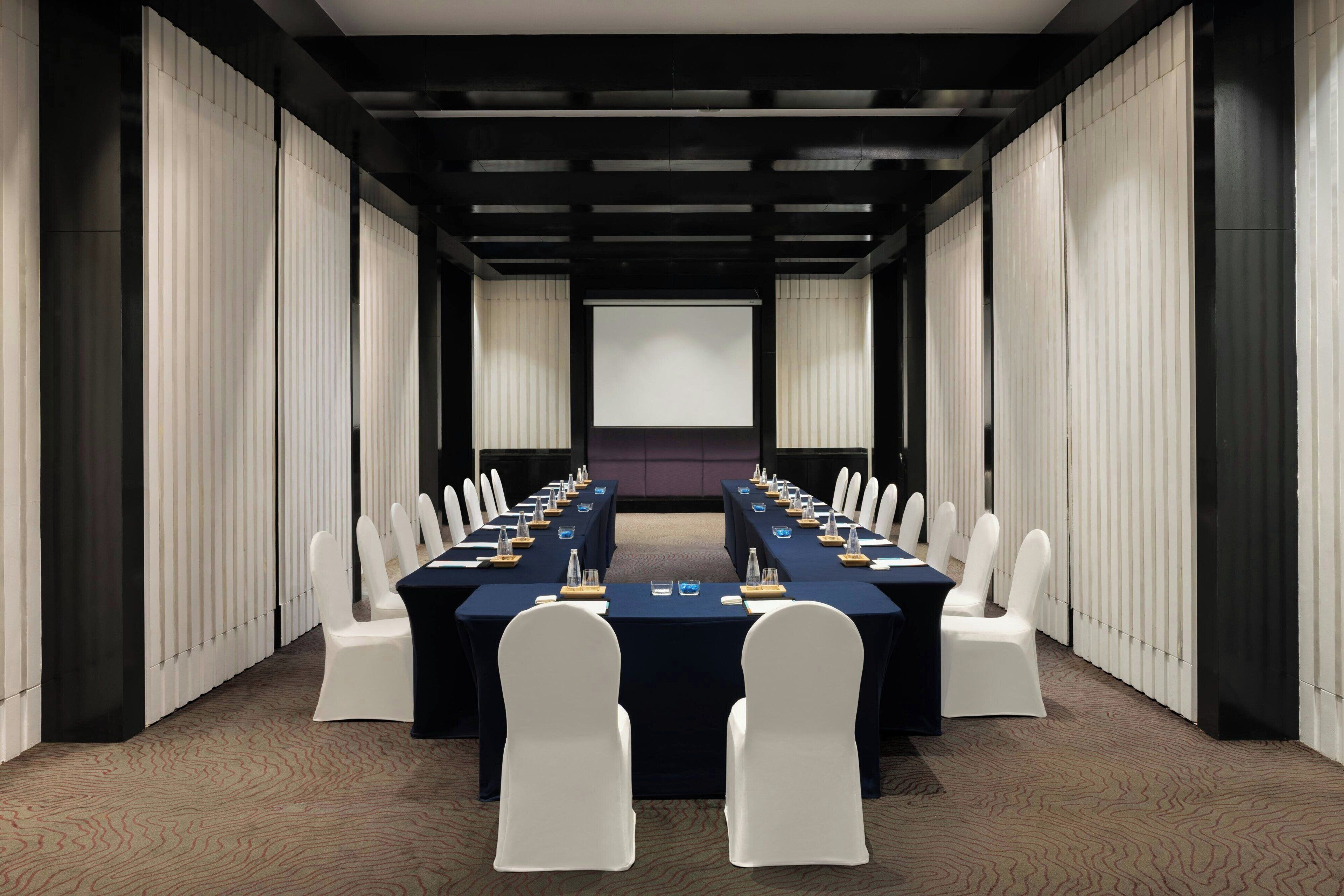 Una sala conferenze con un lungo tavolo, sedie bianche e un grande schermo di fronte.