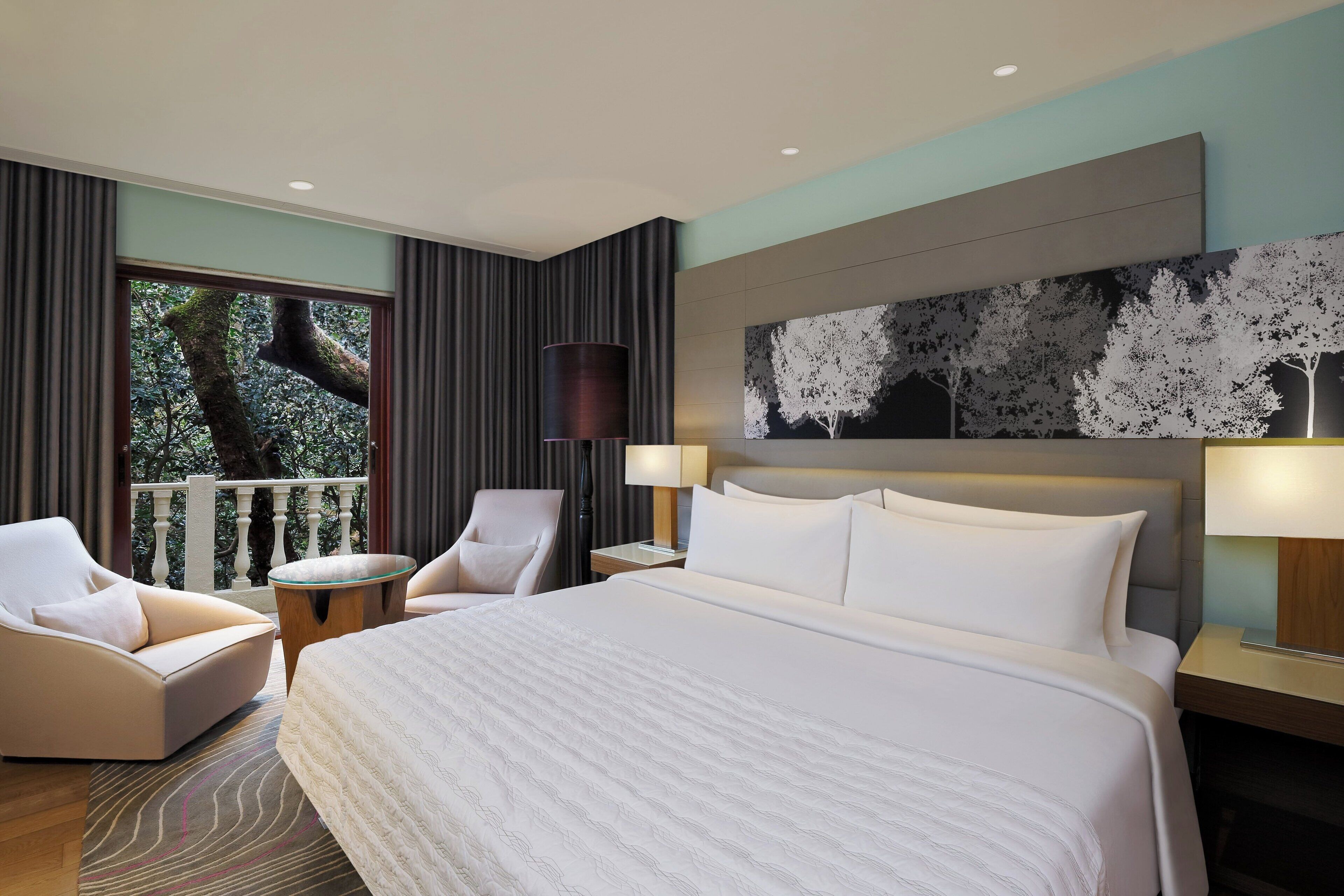 Habitación de hotel con una cama grande, dos sillones, una mesita auxiliar y vistas a árboles por una ventana.