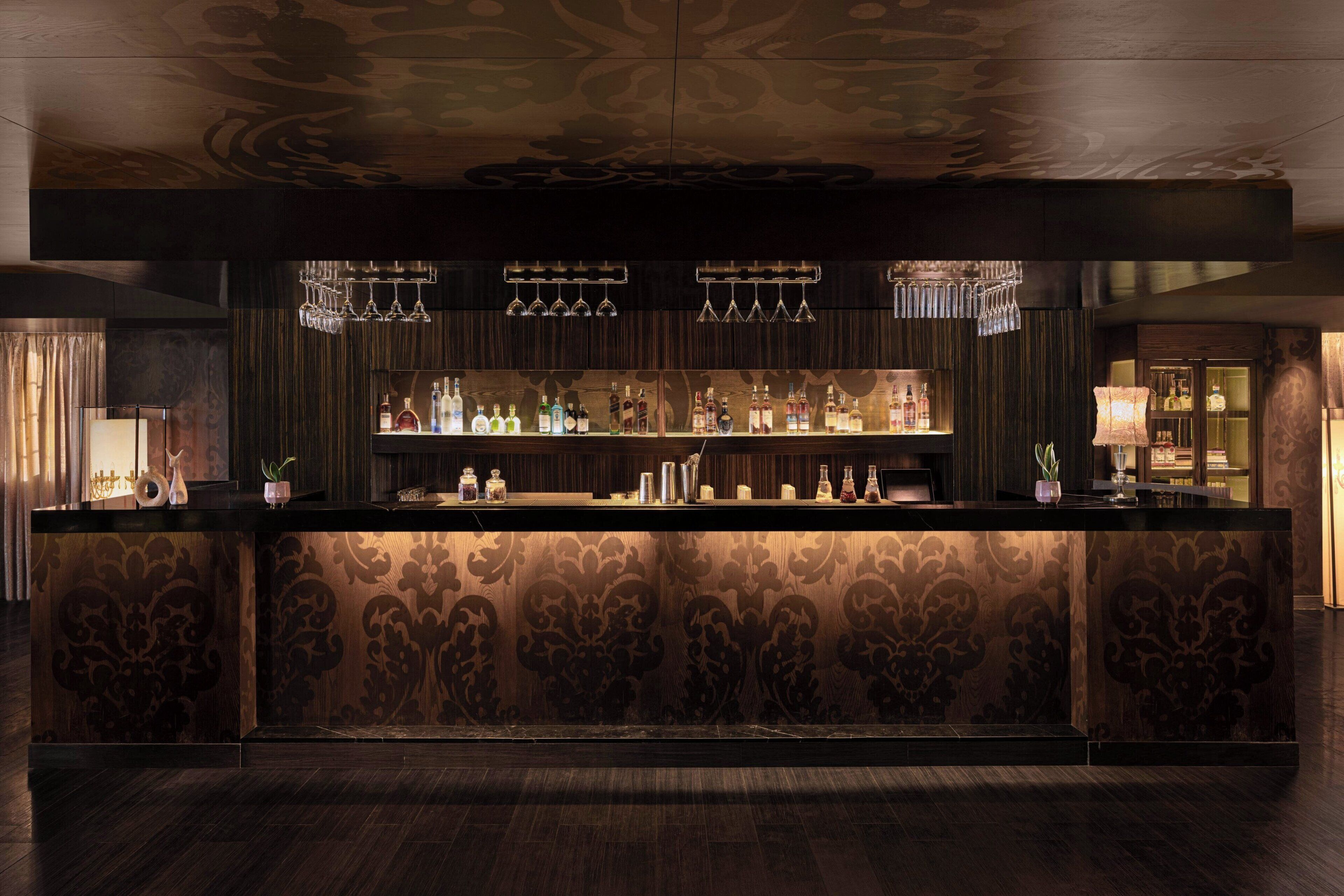 A bar area with a patterned backdrop, shelves with bottles, and hanging glasses.