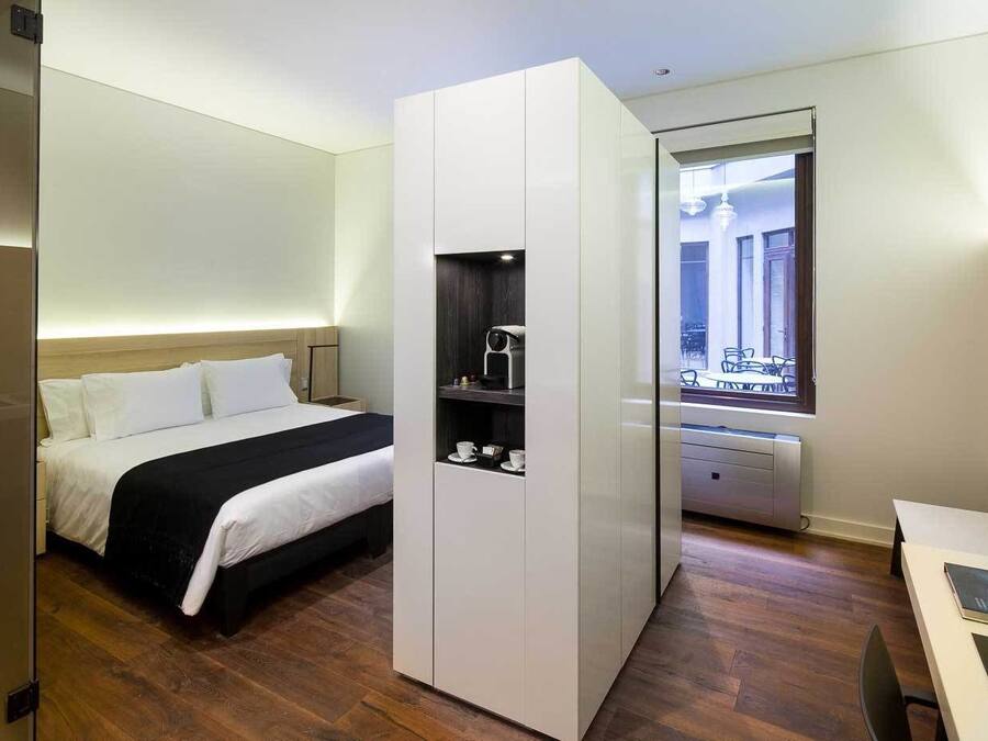 Una habitación de hotel moderna con cama, armario empotrado, kitchenette pequeño y ventana con vistas a la ciudad.