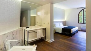 A modern bathroom with a marble wall, a large mirror, and a bathtub. The bedroom features a bed with a black headboard, a bedside table, and a window with a view of greenery. - Altiplanico Bellas Artes (Santiago)