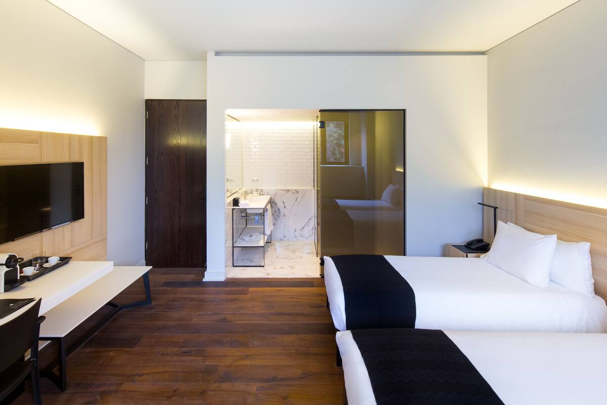 A modern hotel room with a large bed, a desk with a flat-screen TV, a wooden floor, and a bathroom visible through an open door.