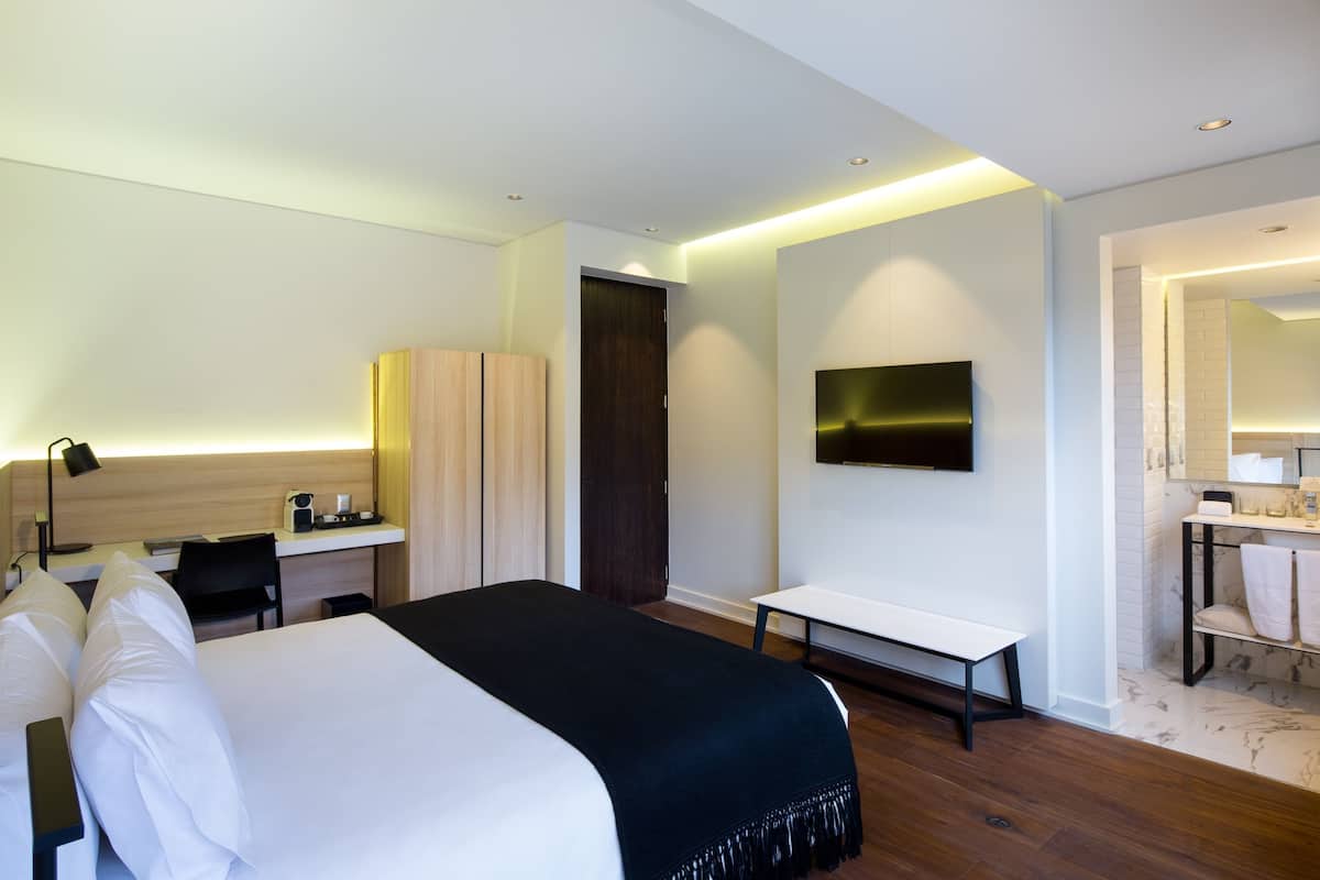 A modern hotel room with a large bed, a desk, a chair, a TV, a bench, and a bathroom with a mirror and towel rack.