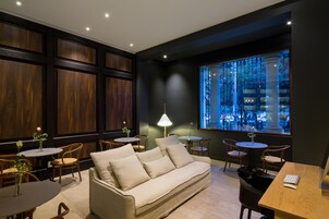 A modern lounge area with a sofa, tables, and chairs, featuring a large window with a city view.