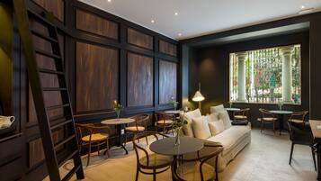 A modern interior with a sofa, round tables, wicker chairs, a ladder, and a large window with a view of trees.