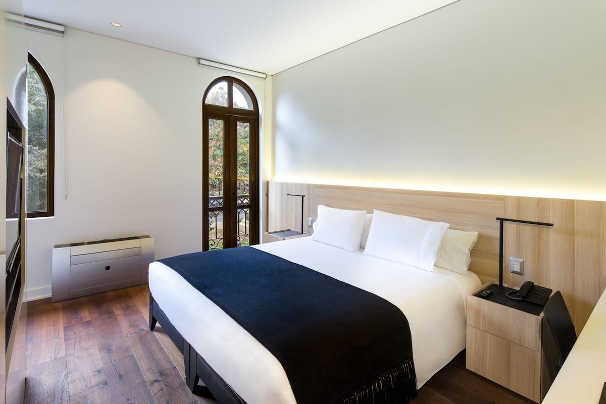 A modern hotel room with a large bed, bedside table, and a view through a window.