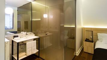 A modern hotel room with a glass-enclosed shower, a marble-topped vanity, a bedside table with a lamp, and a neatly made bed.