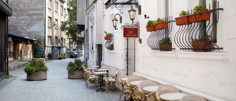 A narrow street with outdoor seating, potted plants, and a building with a sign.