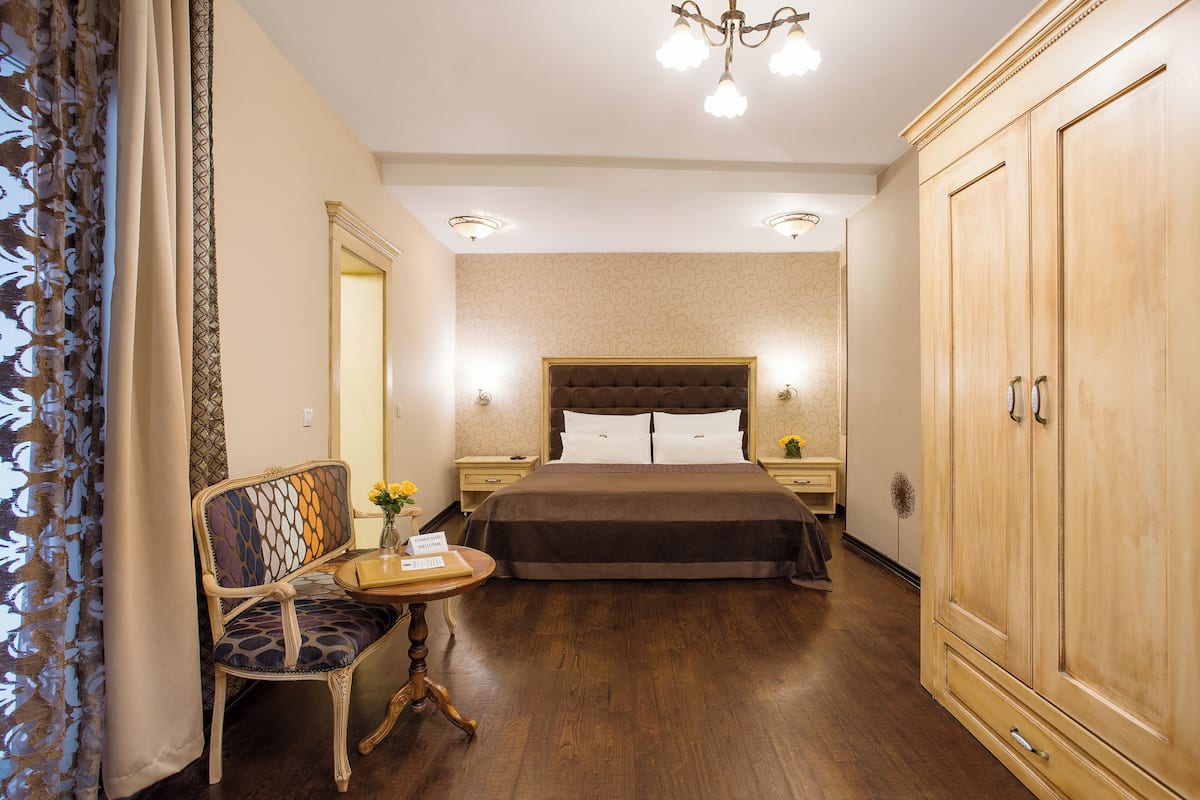 A hotel room with a large bed, a wooden wardrobe, a small table with a vase of flowers, and a chair with a patterned cushion.