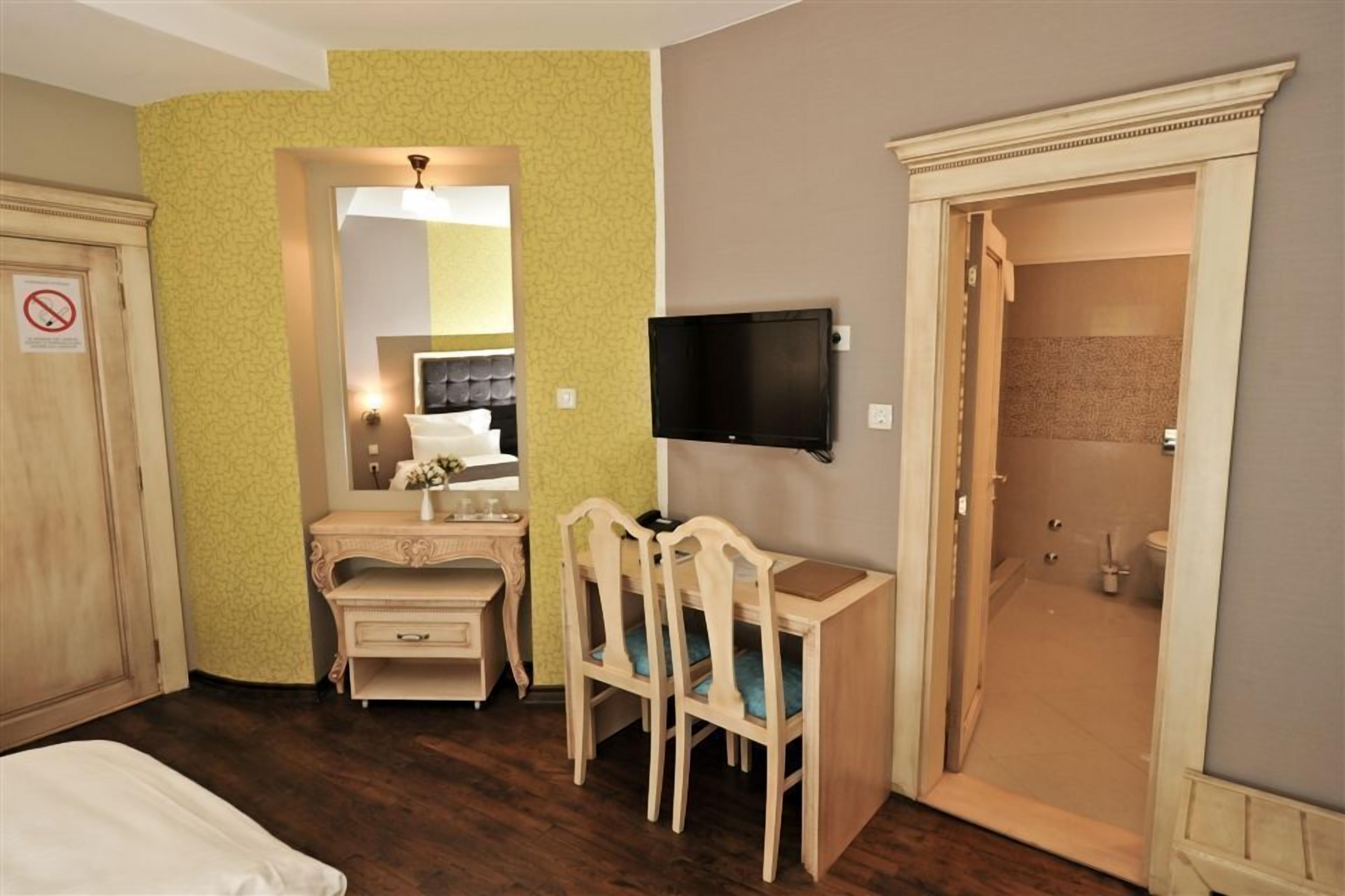 A hotel room with a wooden desk, two chairs, a television, a mirror, and a door leading to a bathroom.