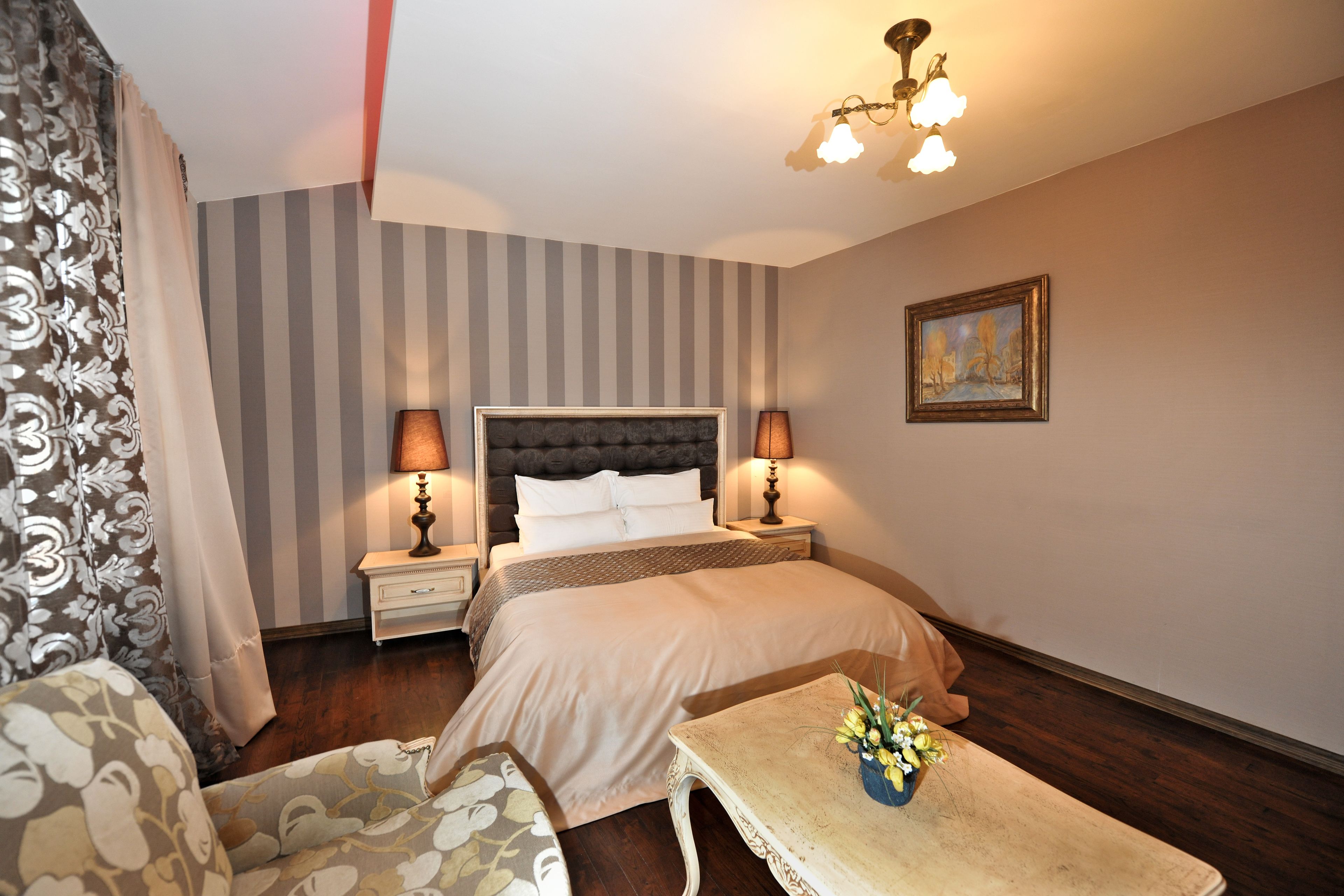 A hotel room with a bed, two bedside tables with lamps, a small table with a vase of flowers, a chair, and a painting on the wall.
