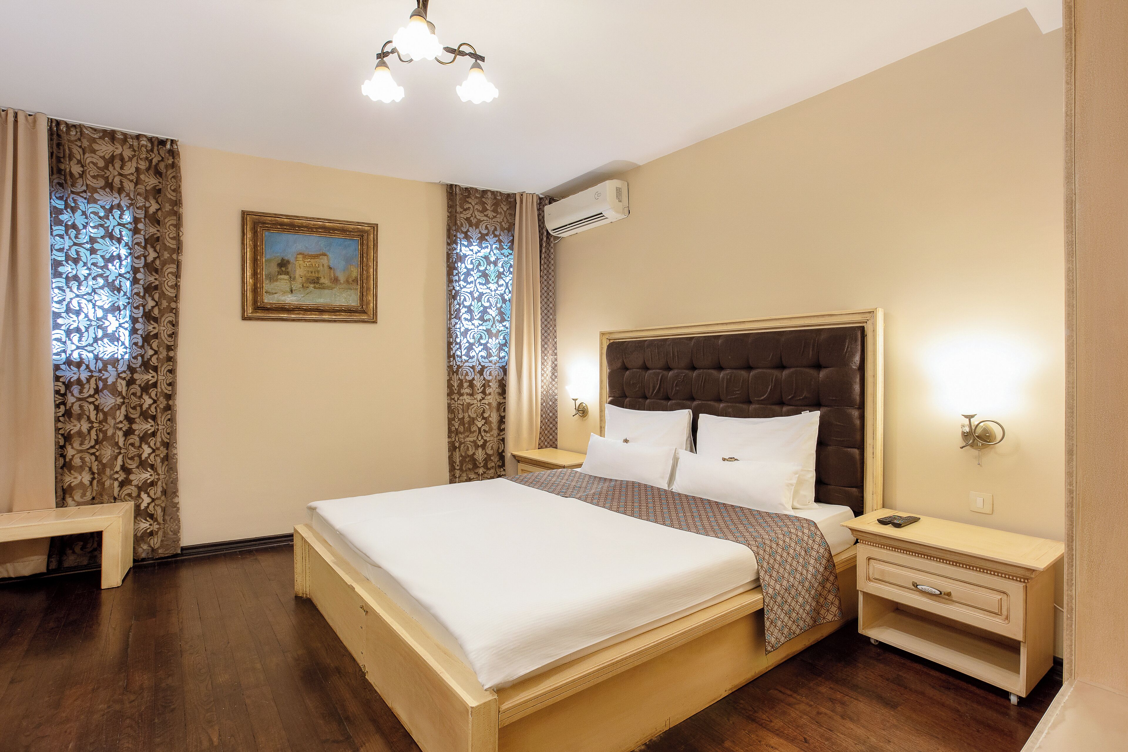 A hotel room with a large bed, a bedside table, a nightstand, an air conditioning unit, and a painting on the wall.