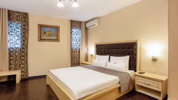 A hotel room with a large bed, a bedside table, a nightstand, an air conditioning unit, and a painting on the wall.