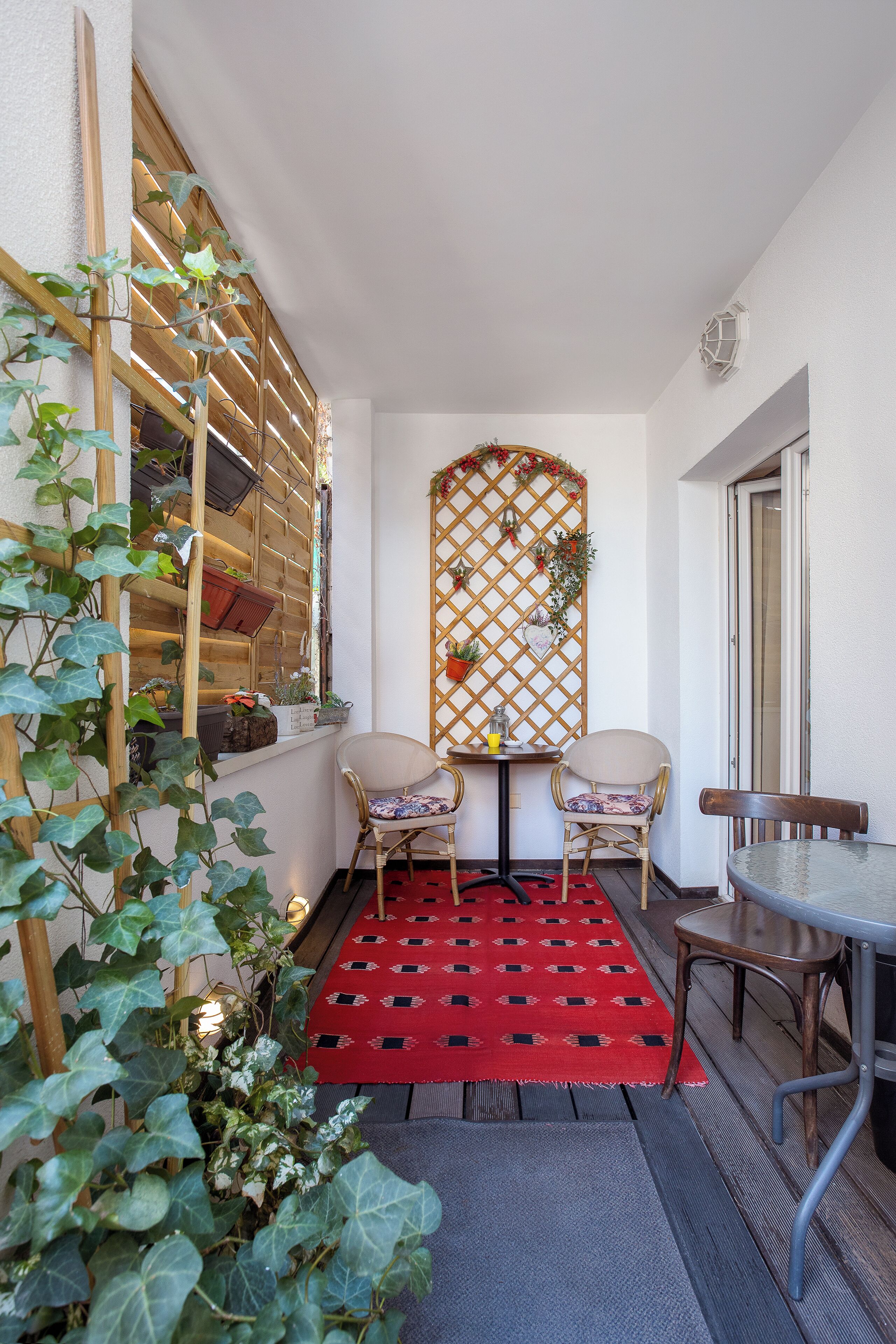 A small patio area with a red patterned rug, two chairs, a table, and a wooden lattice wall with plants.