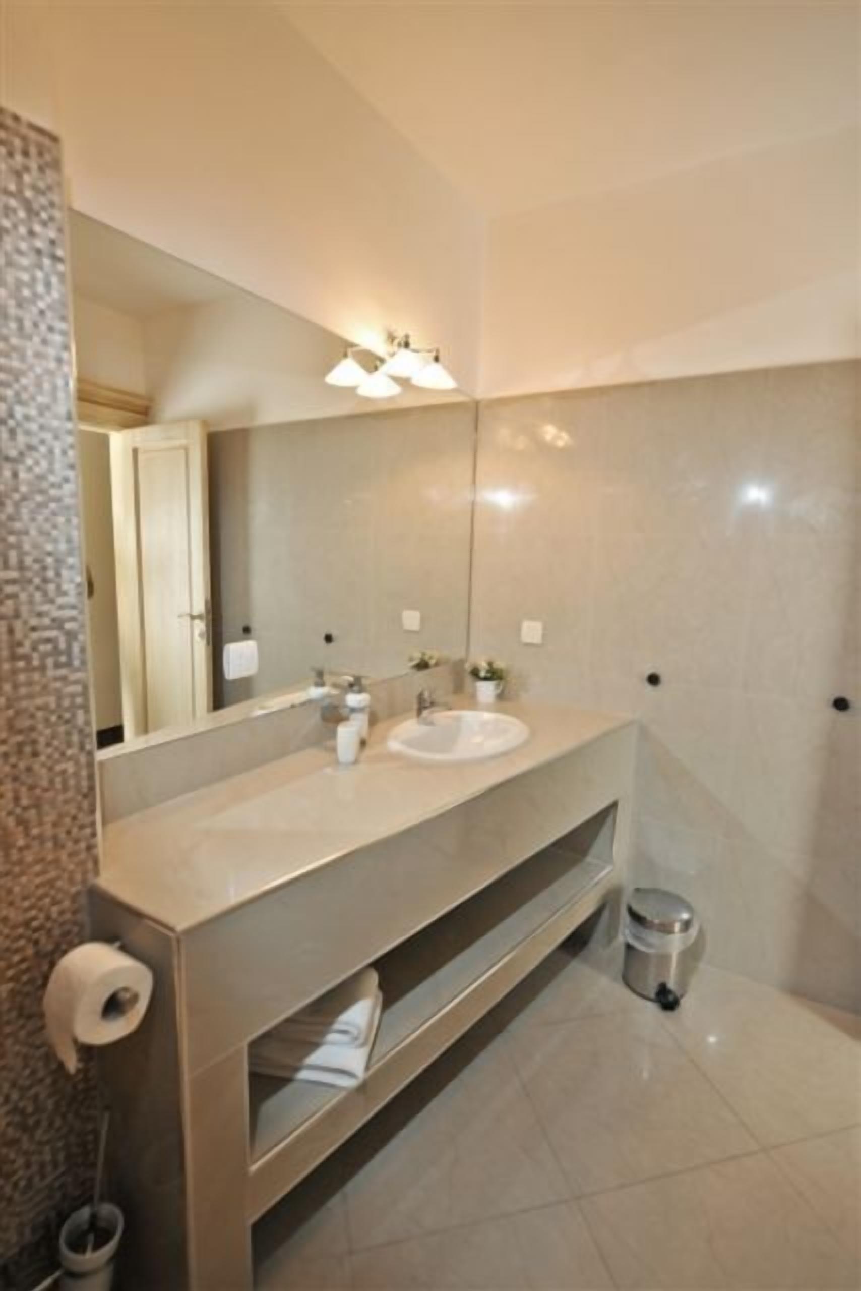 A modern bathroom with a large mirror, two sinks, a towel rack, and a trash bin.