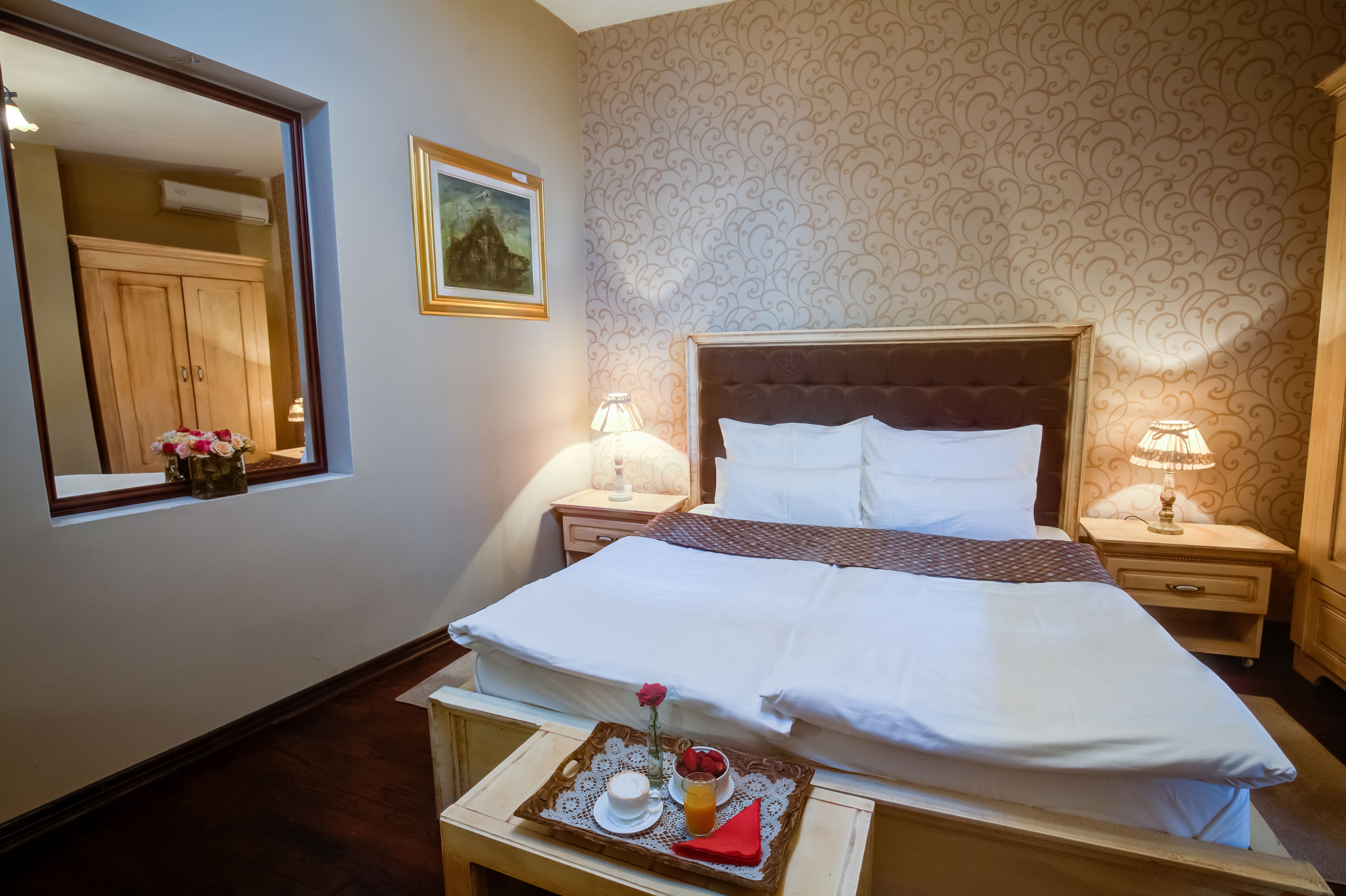 A neatly made bed with white linens, a tray with a cup, a rose, and fruit on a wooden nightstand.