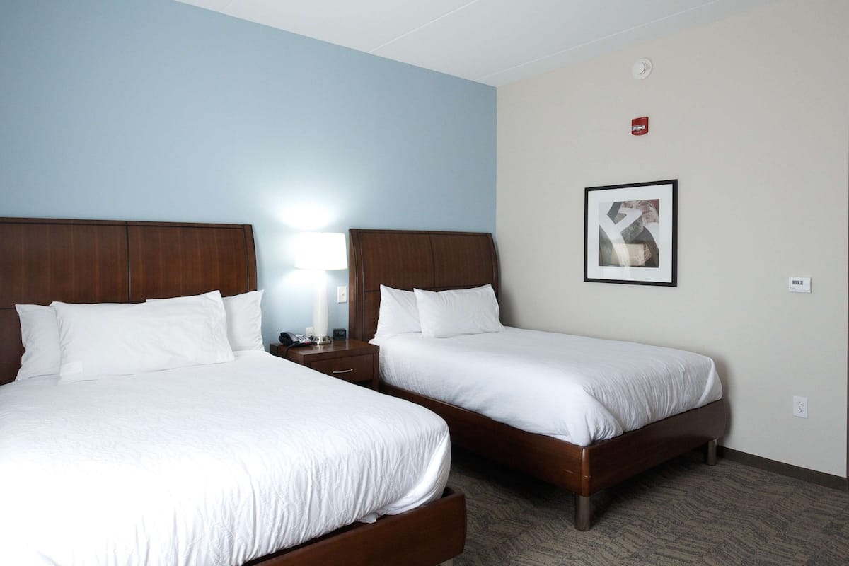 A hotel room with two beds, a nightstand with a lamp, a framed picture on the wall, and a door to the right.