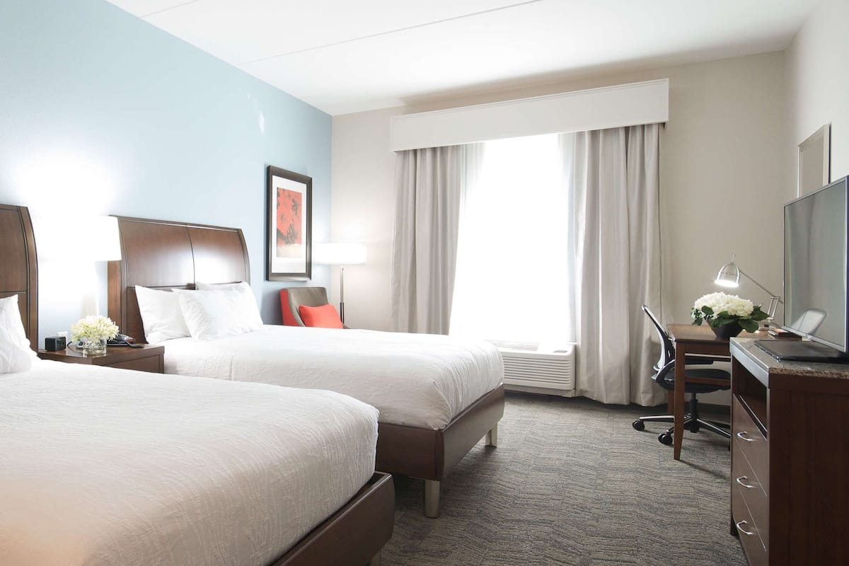 A hotel room with two beds, a desk, a chair, a TV, and a window with curtains.