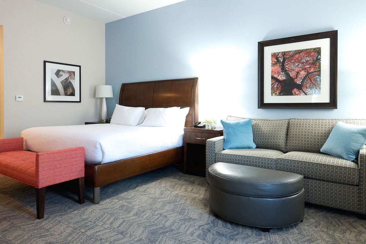 A hotel room with a bed, a red chair, a grey sofa, and a black ottoman. There is a framed picture on the wall and a lamp on the nightstand.