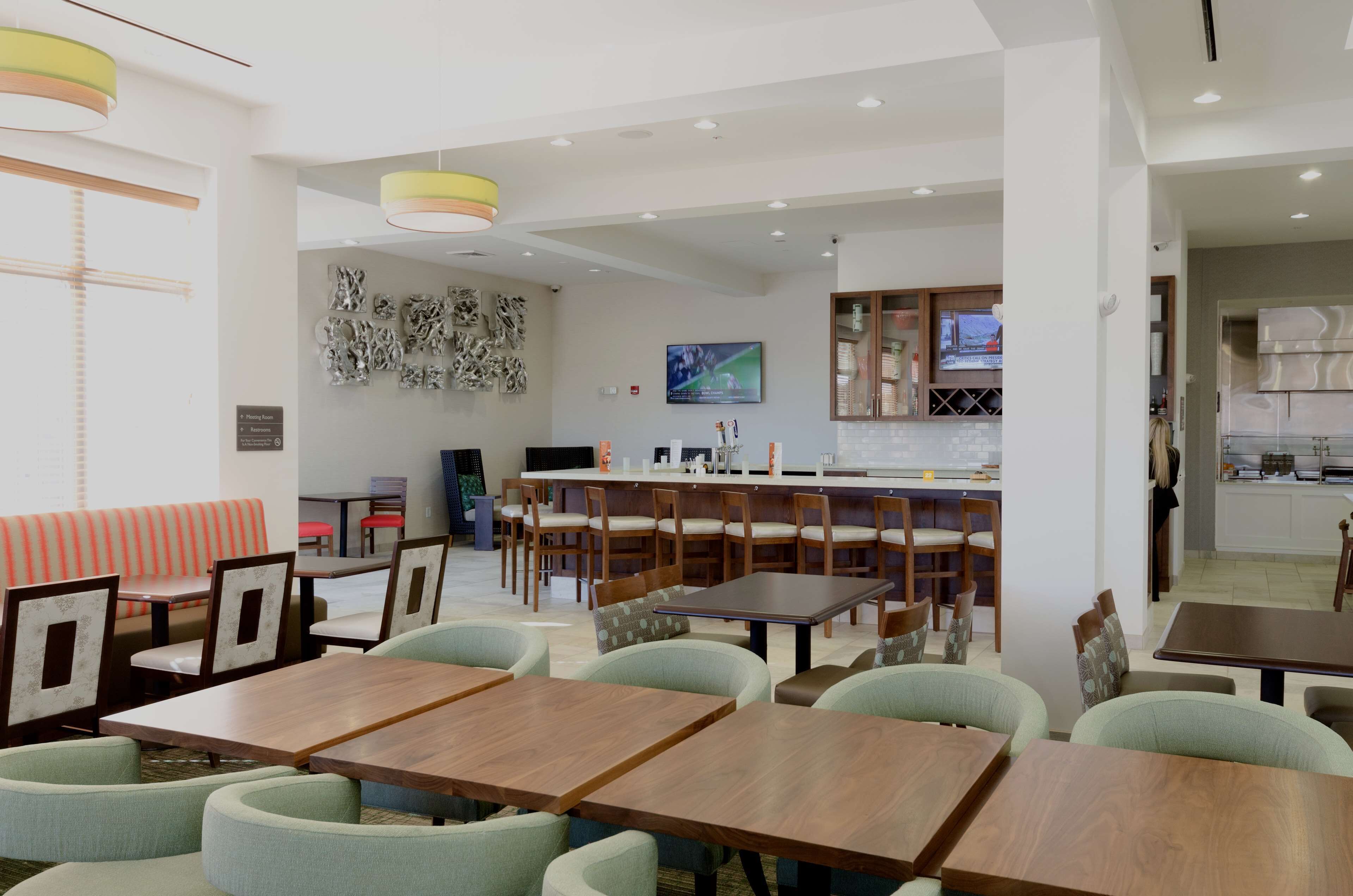 A modern dining area with wooden tables and chairs, a bar area, and a kitchen visible in the background.