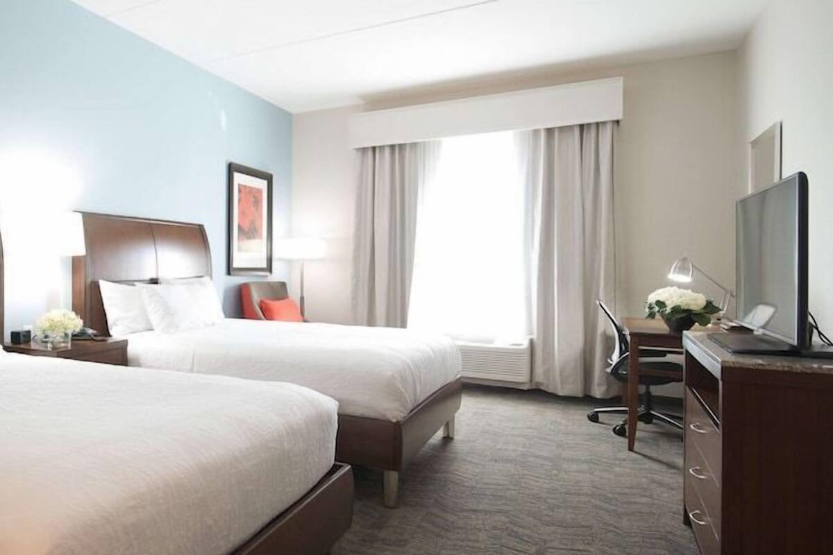 A hotel room with two beds, a desk with a chair, a TV, and a window with curtains.