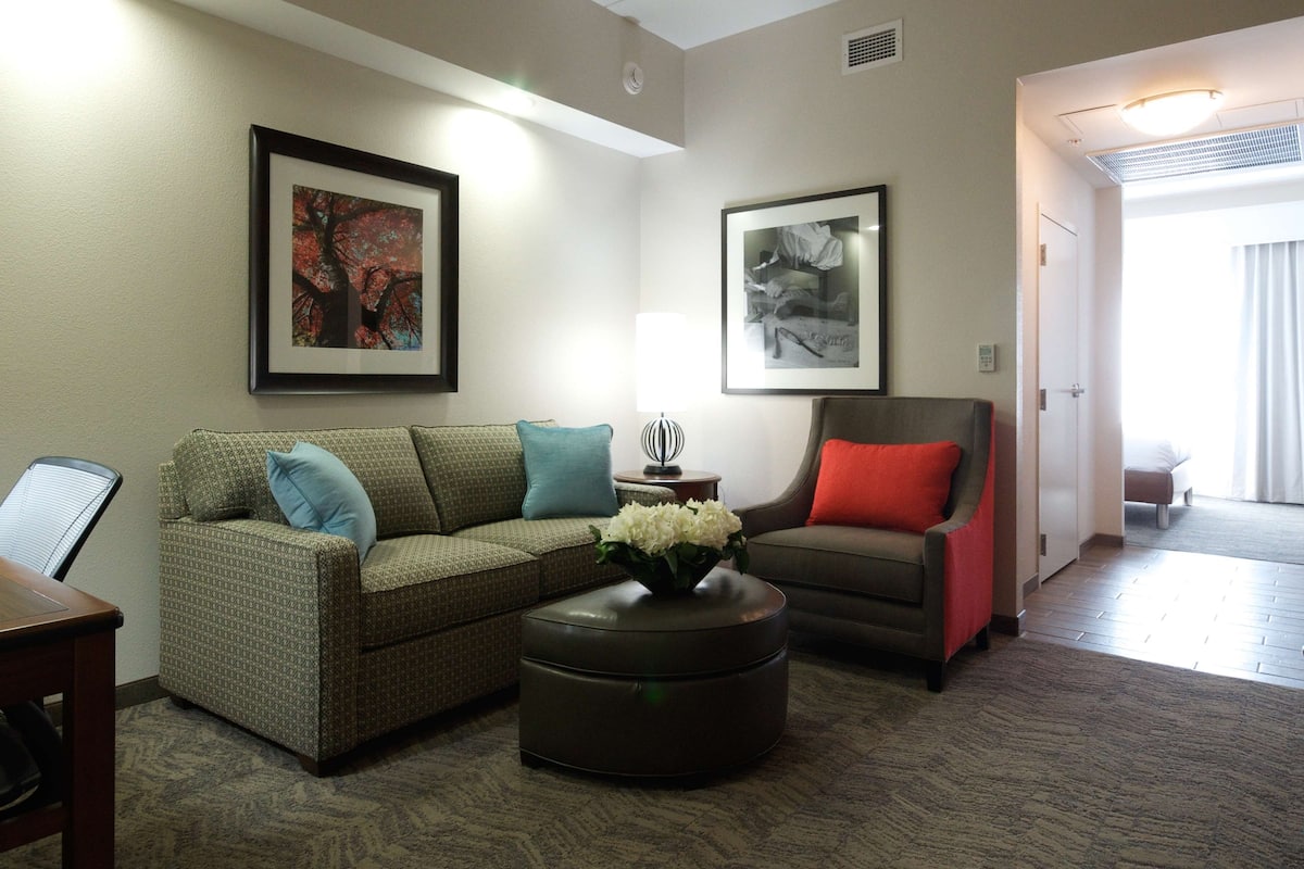A living room with a sofa, armchair, ottoman, and a table. There are framed pictures on the wall.