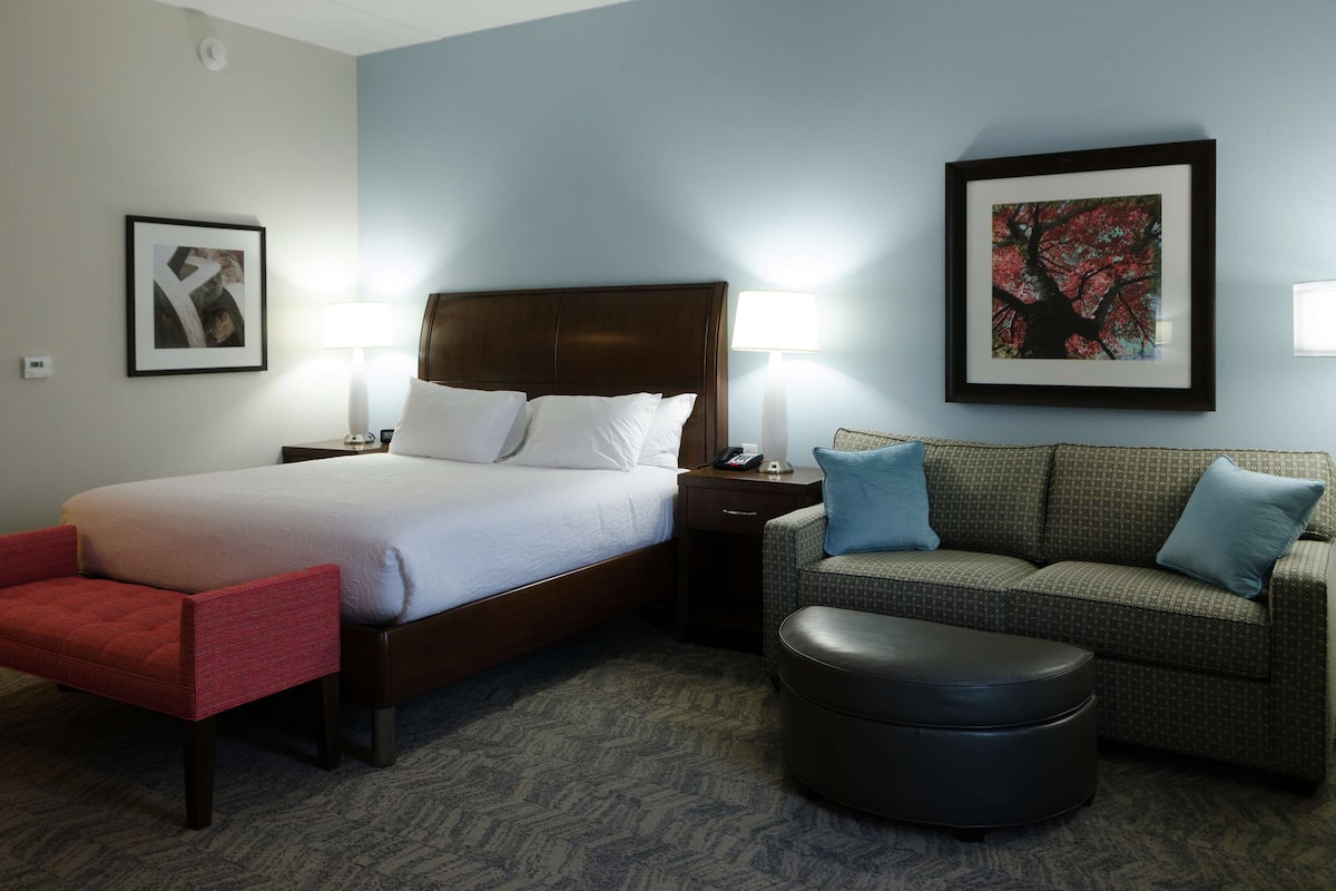 A hotel room with a bed, two bedside tables, a red chair, a patterned sofa, and a black ottoman.