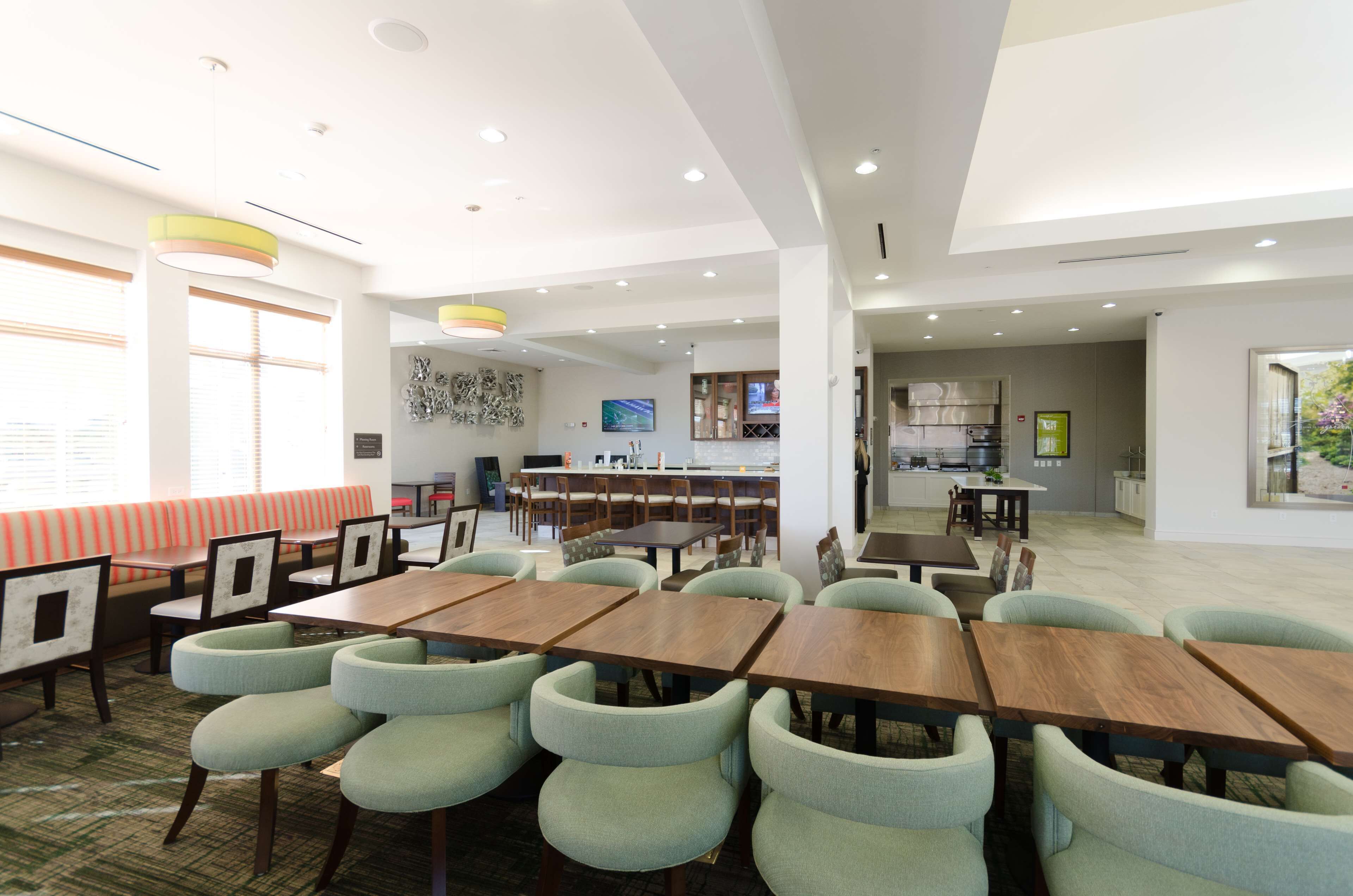 A modern dining area with multiple tables and chairs, a kitchen in the background, and a large window providing natural light.