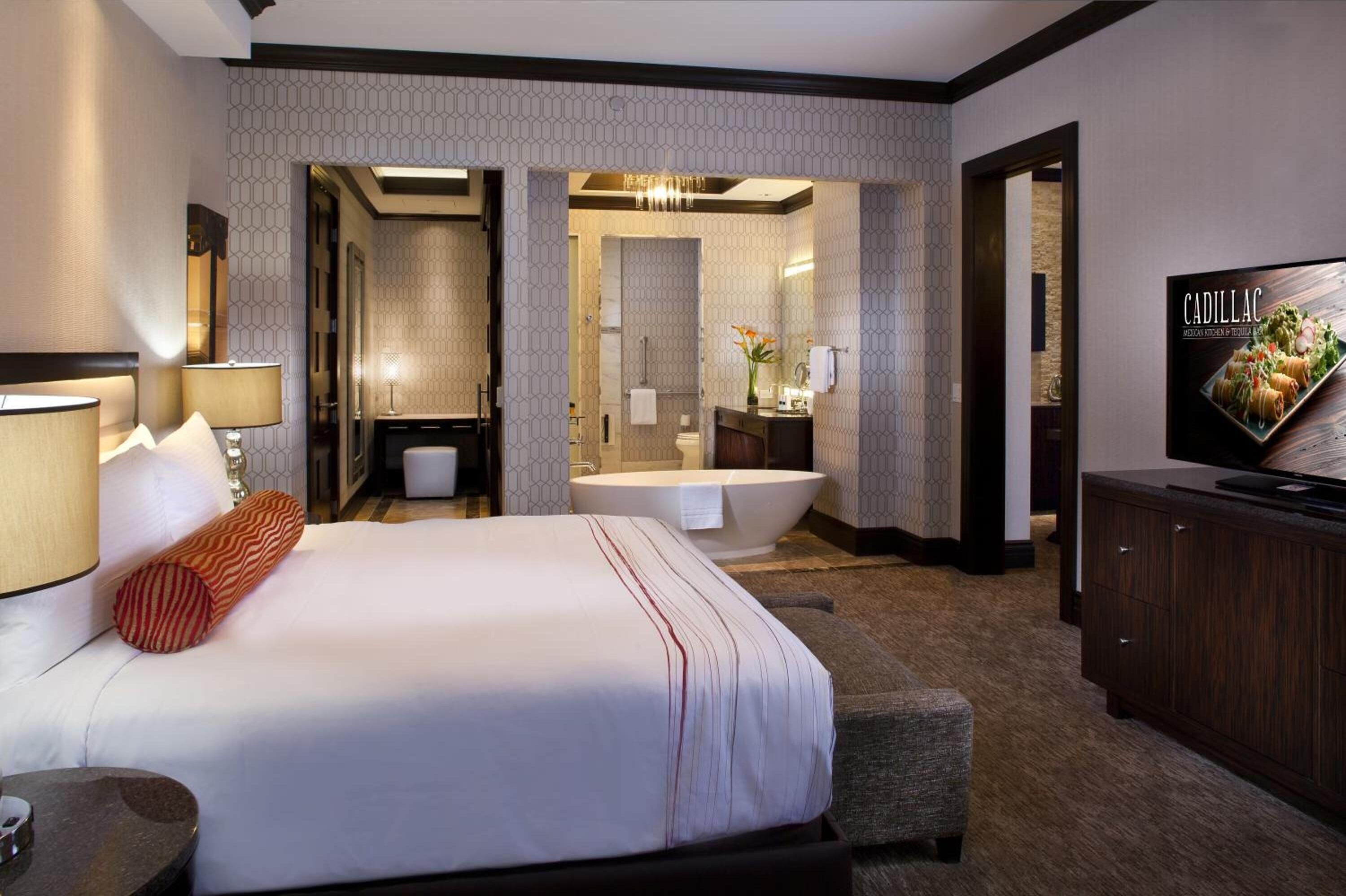 A hotel room with a large bed, a sitting area, a bathtub, a vanity, and a television.
