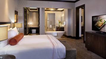 A hotel room with a large bed, a sitting area, a bathtub, a vanity, and a television.